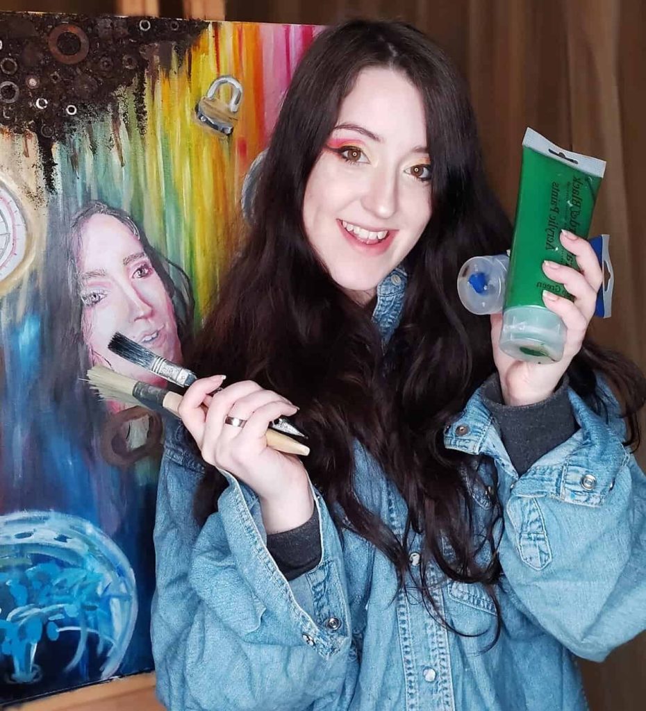 Kirsty MacDonald with a paintbrush and paint with her artwork behind
